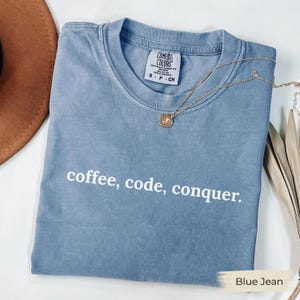 以下が含まれることがあります： 「coffee, code, conquer」の文字が入ったブルージーンズ色のTシャツ。ペンダント付きのゴールドネックレスがシャツの上にかけられています。シャツは折りたたまれており、「Comfort Colors」と書かれたタグが付いています。