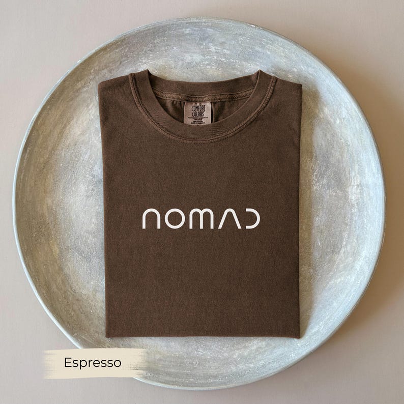 A brown t-shirt with the word "NOMAD" printed on it, laid out on a white plate.