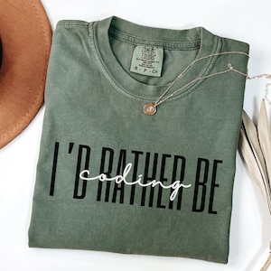 May include: An olive green t-shirt with the phrase "I'd rather be coding" printed in black and white. A gold necklace with a pendant rests on the shirt. The shirt is a Comfort Colors brand, size S-P-CH.