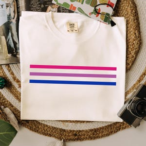 May include: A white t-shirt with three horizontal stripes in pink, purple, and blue, representing the bisexual pride flag. The shirt is a Comfort Colors brand, size XL-TG-EG.