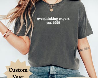 May include: A dark gray t-shirt with the text "overthinking expert est. 1999" in white. The shirt is a crew neck style and is made of a soft material. The bottom of the shirt is visible, with the word "Pepper" printed on the bottom.