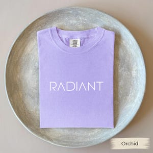 A purple t-shirt with the word "RADIANT" printed on it, laid out on a gray plate.