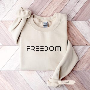 May include: A sand-colored sweatshirt with the word "FREEDOM" printed in black across the front. The sweatshirt is folded neatly on a wooden surface. The sleeves are rolled up, and the overall look is casual and comfortable.