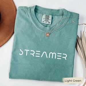 以下が含まれることがあります： 白い文字で「STREAMER」と書かれた、ライトグリーンのTシャツ。Tシャツはクルーネックで半袖です。ペンダント付きのシルバーのネックレスがTシャツの上にかけられています。背景には茶色の帽子が見えます。
