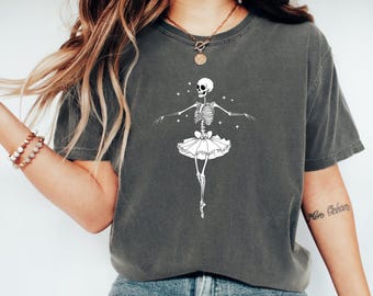 Può includere: T-shirt grigio scuro con un design di ballerina scheletro bianco. Lo scheletro è in posa en pointe, indossa un tutù ed è circondato da stelle. La maglietta ha un girocollo classico e maniche corte.