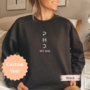 May include: Black sweatshirt with white text reading "PHD" stacked vertically, with "EST 2026" below. The sweatshirt has a crew neck and long sleeves. A peach-colored sticker with the words "Custom Year" is in the lower left corner. The word "Black" is in a white box in the lower right corner.