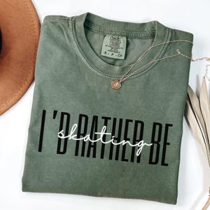 Puede incluir: Una camiseta verde oliva con la frase "I'd rather be skating" en letras blancas y negras. Un collar dorado con un colgante descansa sobre la camiseta. La camiseta es de la marca Comfort Colors.