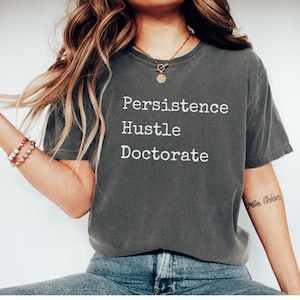 Puede incluir: Camiseta gris oscuro con las palabras "Persistence", "Hustle" y "Doctorate" impresas en blanco. La camiseta es de cuello redondo y está hecha de un material suave. La camiseta se usa con vaqueros azules.