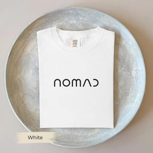 A white t-shirt with the word "NOMAD" printed on it, laid out on a gray plate.