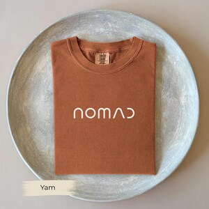 a brown t-shirt with the word "NOMAD" printed on it, placed on a gray plate.