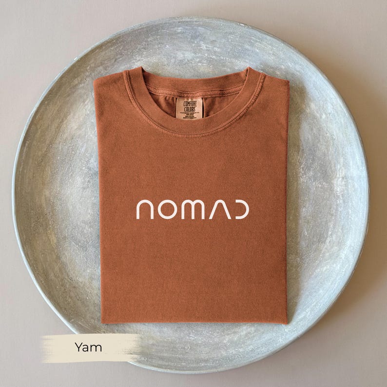 a brown t-shirt with the word "NOMAD" printed on it, placed on a gray plate.