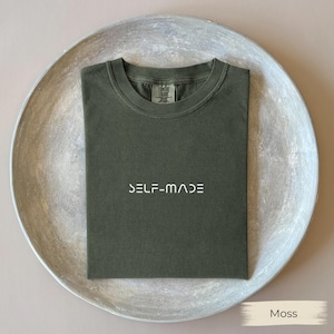 May include: A moss green t-shirt with the word "SELF-MADE" printed in white. The shirt is folded neatly on a decorative, round, silver-colored tray. The shirt has a crew neck and short sleeves. The tag on the shirt reads "COMFORT COLORS".