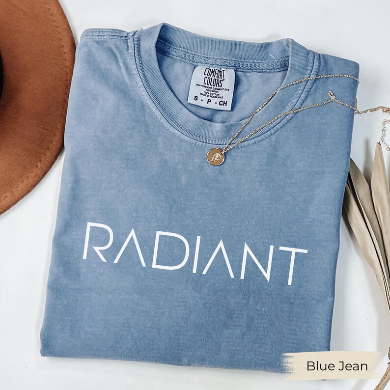 A blue t-shirt with the word "RADIANT" printed on it, along with a necklace and a brown hat, all laid out on a white surface.