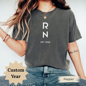 May include: A dark gray t-shirt with the letters "RN" in white, stacked vertically, and the text "EST 2026" below. The shirt is a solid color and has a crew neck.
