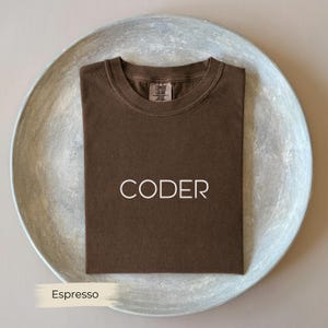 Może przedstawiać: Koszulka w kolorze espresso z napisem "CODER" białymi literami. Koszulka jest starannie złożona na okrągłej, jasnoszarej tacy. Koszulka ma okrągły dekolt i metkę "Comfort Colors".