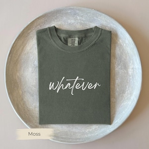 May include: A moss green t-shirt with the word "whatever" in white script. The shirt is folded neatly on a silver-colored, round tray. The shirt has a crew neck and short sleeves.