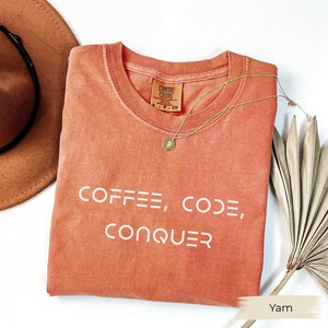 以下が含まれることがあります： 「COFFEE, CODE, CONQUER」の白いブロック文字がプリントされた、ヤム色のComfort Colors Tシャツ。ペンダント付きのゴールドネックレスがシャツの上にあります。茶色の帽子と乾燥した植物が背景にあります。
