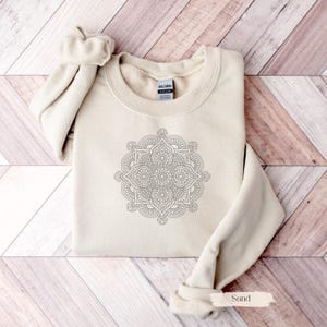 May include: A sand-colored sweatshirt with a crew neck and long sleeves. A detailed black and white mandala design is centered on the front. The word "Sand" is printed on a small label at the bottom.