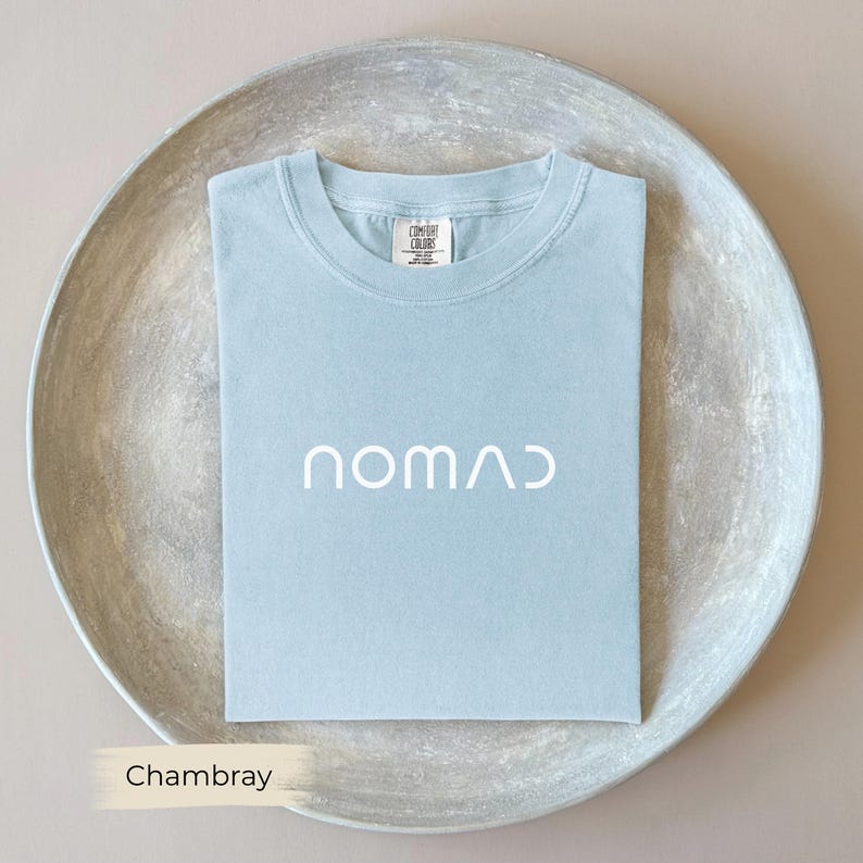 A light blue t-shirt with the word "NOMAD" printed on it, laid out on a round, gray plate.