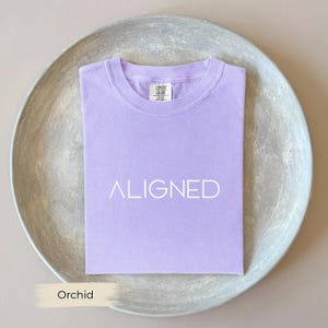 Könnte beinhalten: Ein gefaltetes, orchideenfarbenes T-Shirt mit dem weißen Aufdruck "ALIGNED". Das Shirt wird auf einem silberfarbenen, runden Tablett präsentiert. Das Wort "Orchid" ist in einem cremefarbenen Rechteck unten aufgedruckt.