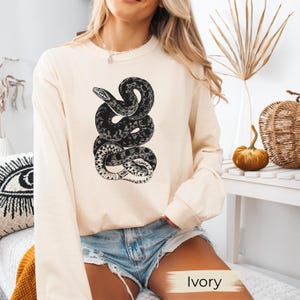 Op de afbeelding: Ivoor kleurige longsleeve shirt met een gedetailleerde zwart-wit illustratie van een opgerolde slang. Het slang ontwerp is gecentreerd op de voorkant van het shirt. Het shirt heeft een neutrale kleur.