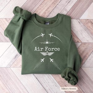 Op de afbeelding: Militairgroen sweatshirt met witte "Air Force" tekst en vliegtuigafbeeldingen. Het ontwerp bevat een ster- en vleugelembleem. De sweatshirt is gemaakt van een zacht materiaal.