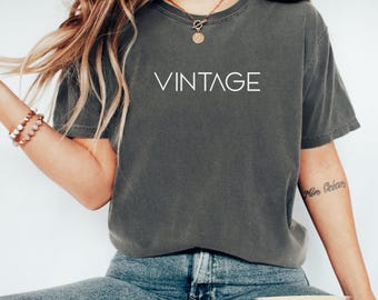Puede incluir: Una camiseta gris oscuro con la palabra "VINTAGE" impresa en blanco. La camiseta es de color liso y tiene mangas cortas. La modelo lleva vaqueros azules.