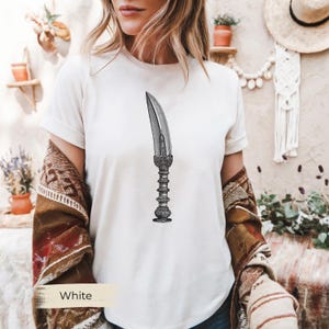 May include: White t-shirt featuring a detailed black and white illustration of a dagger. The dagger has an ornate handle and a curved blade. The word "White" is printed on a beige rectangle at the bottom.