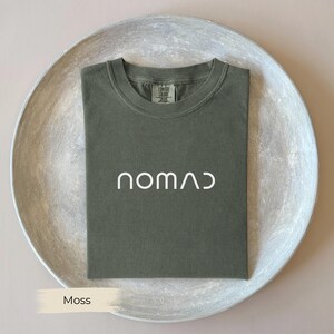 A green t-shirt with the word "NOMAD" printed on it, laid out on a white plate.