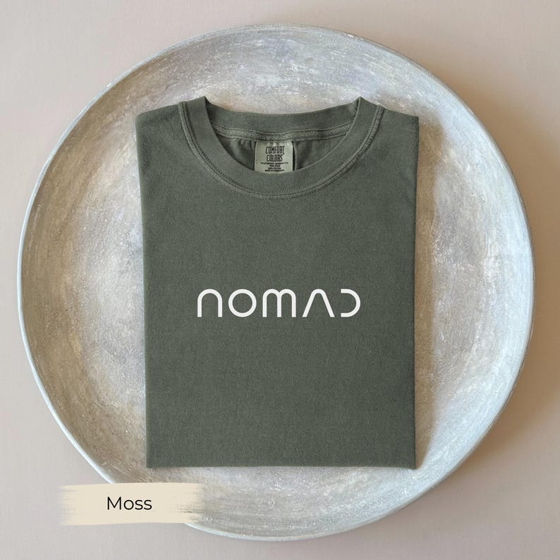 A green t-shirt with the word "NOMAD" printed on it, laid out on a white plate.