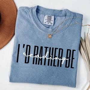 Op de afbeelding: Een blauw Comfort Colors T-shirt met de tekst "I'd rather be reading" in zwart en wit. Een gouden ketting met een hanger is over het shirt gedrapeerd. Het shirt is op een wit oppervlak gevouwen.