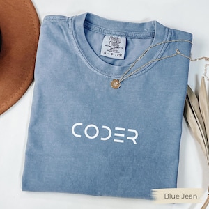May include: A blue jean colored t-shirt with the word "CODER" printed in white. A gold necklace with a pendant is draped across the shirt. The shirt is folded and has a tag that says "Comfort Colors".
