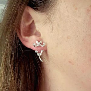 May include: Silver cat-shaped earrings. The earrings feature a cat design with outstretched paws and a tail. The cat's head has a small, red gem. The earrings are worn on the ear.