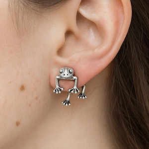 Könnte beinhalten: Silberne Frosch-Ohrringe. Die Ohrringe haben ein Frosch-Design, wobei sich der Körper des Frosches auf der Vorderseite des Ohrs und die Beine hinter dem Ohr befinden. Der Frosch hat große Augen und detaillierte Füße. Die Ohrringe sind aus glänzendem Metall.