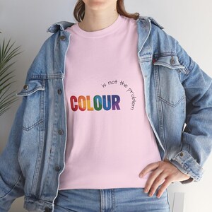 Könnte beinhalten: Hellrosa T-Shirt mit dem Text "COLOUR is not the problem" in Regenbogenfarben. Die Person trägt eine hellblaue Jeansjacke über dem T-Shirt.
