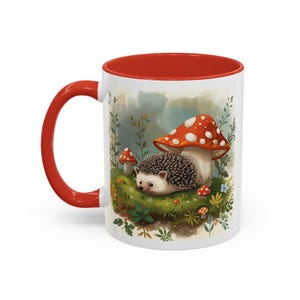 Könnte beinhalten: Weiße Keramiktasse mit rotem Griff und Innenraum. Die Tasse zeigt eine skurrile Illustration eines Igels inmitten von Pilzen und Grün. Das Design umfasst einen großen Pilz mit roten und weißen Punkten.