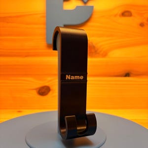 Puede incluir: Un soporte para auriculares de metal negro con una parte superior curva y una base de dos puntas. El soporte presenta una banda horizontal en el centro con la palabra Name grabada en una fuente de color claro.