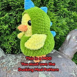 May include: A plush, crocheted duckling wearing a green dinosaur costume with blue spikes. The duckling has a yellow body, orange beak, and black eyes. Text on the image reads: "This is a MOD. You must own the Ducking Mad Pattern."