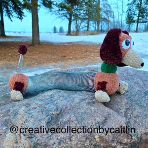 May include: A crocheted Slinky Dog toy, featuring a brown and tan head, ears, and body with a spring-like midsection. The toy has large, blue eyes and a green collar. The toy is resting on a large rock. The text "@creativecollectionbycaitlin" is at the bottom.
