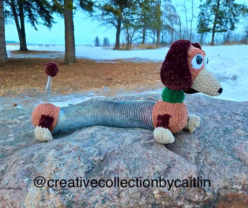 May include: A crocheted Slinky Dog toy, featuring a brown and tan head, ears, and body with a spring-like midsection. The toy has large, blue eyes and a green collar. The toy is resting on a large rock. The text "@creativecollectionbycaitlin" is at the bottom.