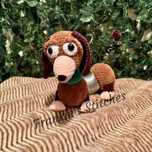 May include: A handmade crochet dog toy in brown, tan, and burgundy. The dog has large white eyes, a pink nose, and a green collar. The dog's body is connected to a silver slinky-like coil. The text "Frankie's Stitches" is at the bottom.