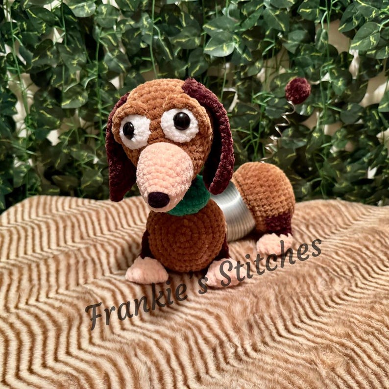 May include: A handmade crochet dog toy in brown, tan, and burgundy. The dog has large white eyes, a pink nose, and a green collar. The dog's body is connected to a silver slinky-like coil. The text "Frankie's Stitches" is at the bottom.