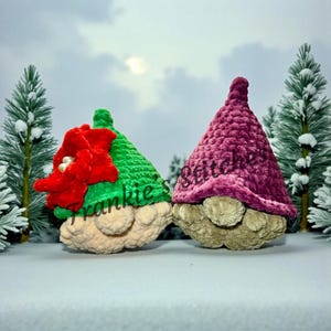 May include: Two plush gnome figures with conical hats. One gnome has a green hat with a red flower and the other has a purple hat. The gnomes have tan faces and bodies. The background is a winter scene with snow and evergreen trees.