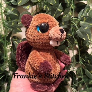 May include: A handmade brown and burgundy plush beaver toy with large, blue-accented eyes and a white tooth. The plushie has a soft, textured appearance and the text "Frankie's Stitches" is visible on the bottom. The beaver is set against a backdrop of green leaves.