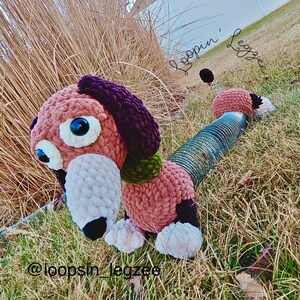 May include: A handmade crochet dachshund dog toy in shades of pink, purple, and black. The dog has large, expressive eyes and a long, tubular body. The text "Loopin' Legzzee" and "@loopsin_legzzee" are visible.