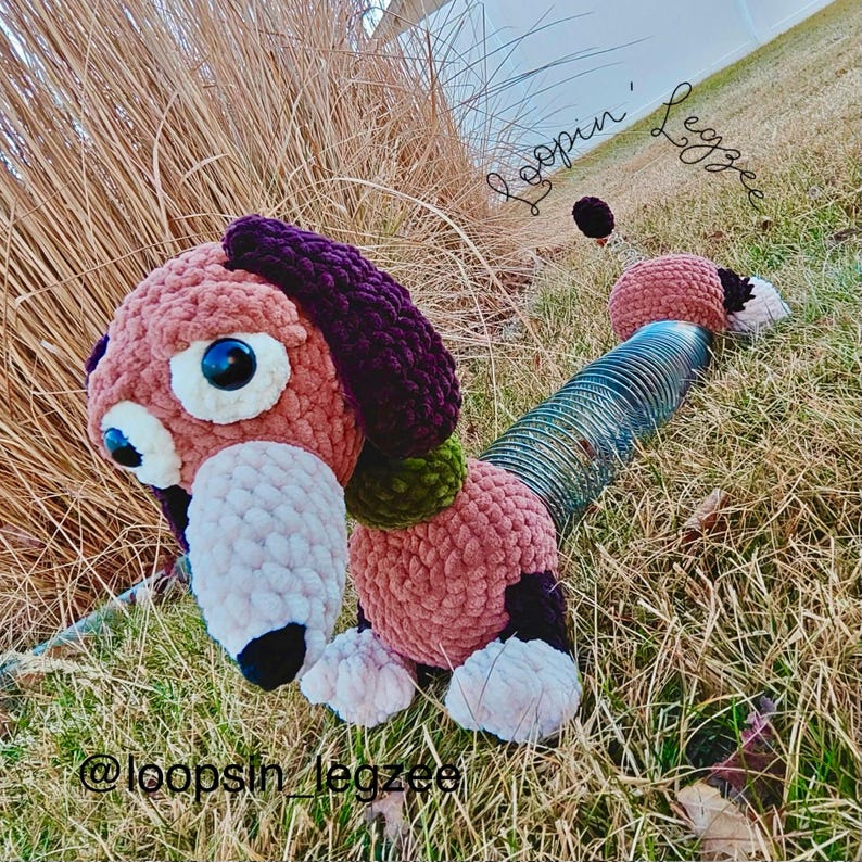 May include: A handmade crochet dachshund dog toy in shades of pink, purple, and black. The dog has large, expressive eyes and a long, tubular body. The text "Loopin' Legzzee" and "@loopsin_legzzee" are visible.