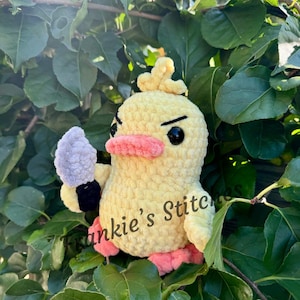 Puede incluir: Un pato amarillo de ganchillo hecho a mano con una expresi&oacute;n de mal humor, ojos negros y un pico y patas rosas. Sostiene un cuchillo de ganchillo gris y tiene el texto "Frankie's Stitches" en el lateral. El pato est&aacute; rodeado de hojas verdes.