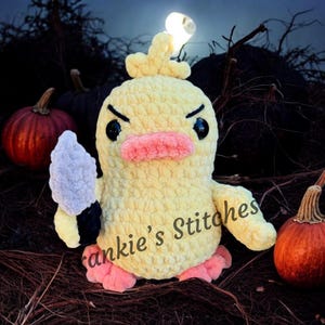 Puede incluir: Un pato de ganchillo amarillo hecho a mano con ojos negros, un pico rosa y pies rosas, sosteniendo un cuchillo gris y negro. El pato tiene una expresi&oacute;n de enfado. El texto "Frankie's Stitches" est&aacute; en el pato. Las calabazas est&aacute;n en el fondo.