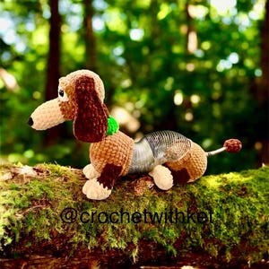 May include: A crocheted dachshund dog toy with a brown and tan body, floppy ears, and a spring-like midsection. The toy has a green collar and a small, round tail. The background is a blurred forest scene.