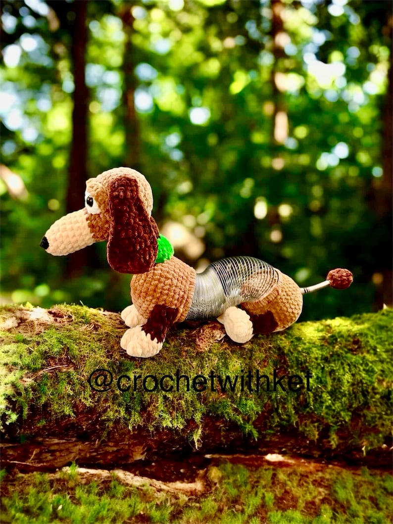 May include: A crocheted dachshund dog toy with a brown and tan body, floppy ears, and a spring-like midsection. The toy has a green collar and a small, round tail. The background is a blurred forest scene.